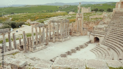 Ruins of Roman theatre in ancient settlement of Dougga in Tunisia with semi-circular orchestra, rows of stone seating and stage columns surrounded by vibrant spring greenery under cloudy sky. High