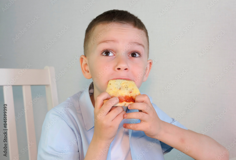 Happy little boy enjoying lunch eating a piece of pizza at home in the kitchen. Children love junk food. Baby food concept. Selective focus. Happy little boy enjoying lunch eating a piece of pizza at home in the kitchen. Children love junk food. Baby food concept. Selective focus.