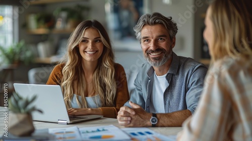 A professional financial planner sitting at a white office desk, explaining investment options to a young couple. The table is organized with a laptop, graphs, and financial reports. The image is