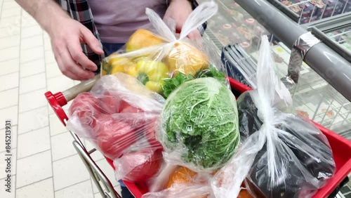 Man in a supermarket. Man Young European Buyer Positive Bearded Long Haired, With Basket In Hands, Shopping Choosing Vegetables Organic In Supermarket.