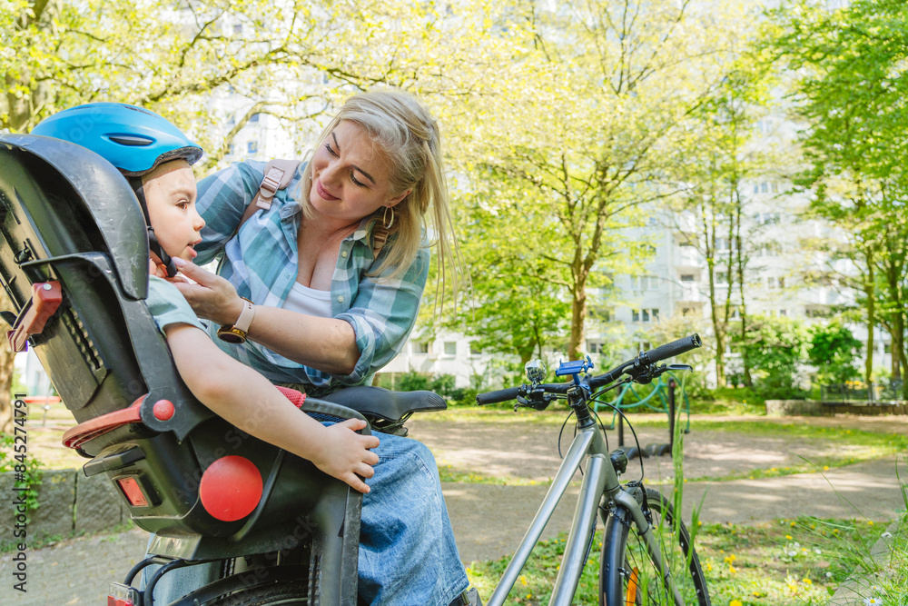 Mother fastening cycling helmet of son sitting on bicycle seat in park Mother fastening cycling helmet of son sitting on bicycle seat in park