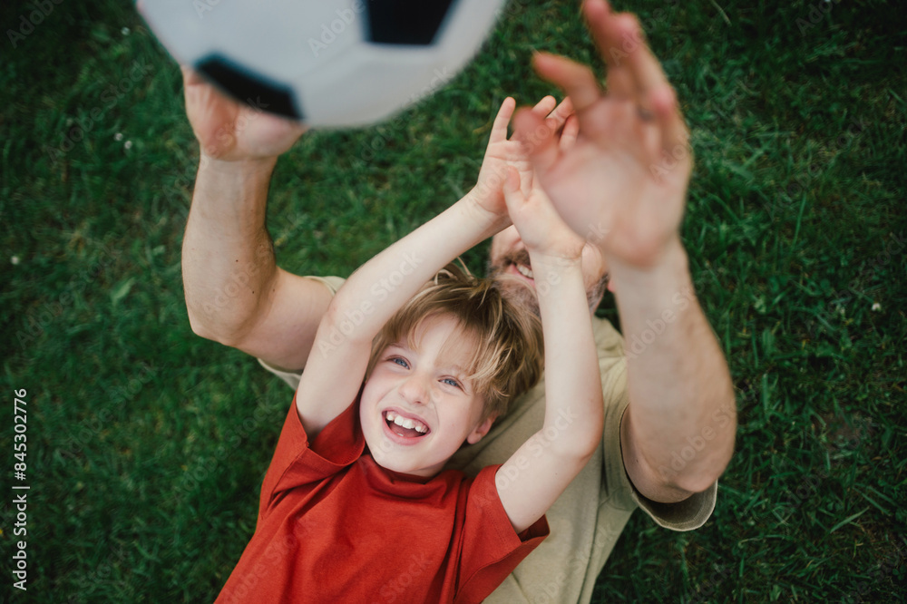 Obraz premium Dad lying on grass with son, laughing and having fun. Father and boy playing football, catching each other, falling to the ground. Fatherhood and Father's Day.