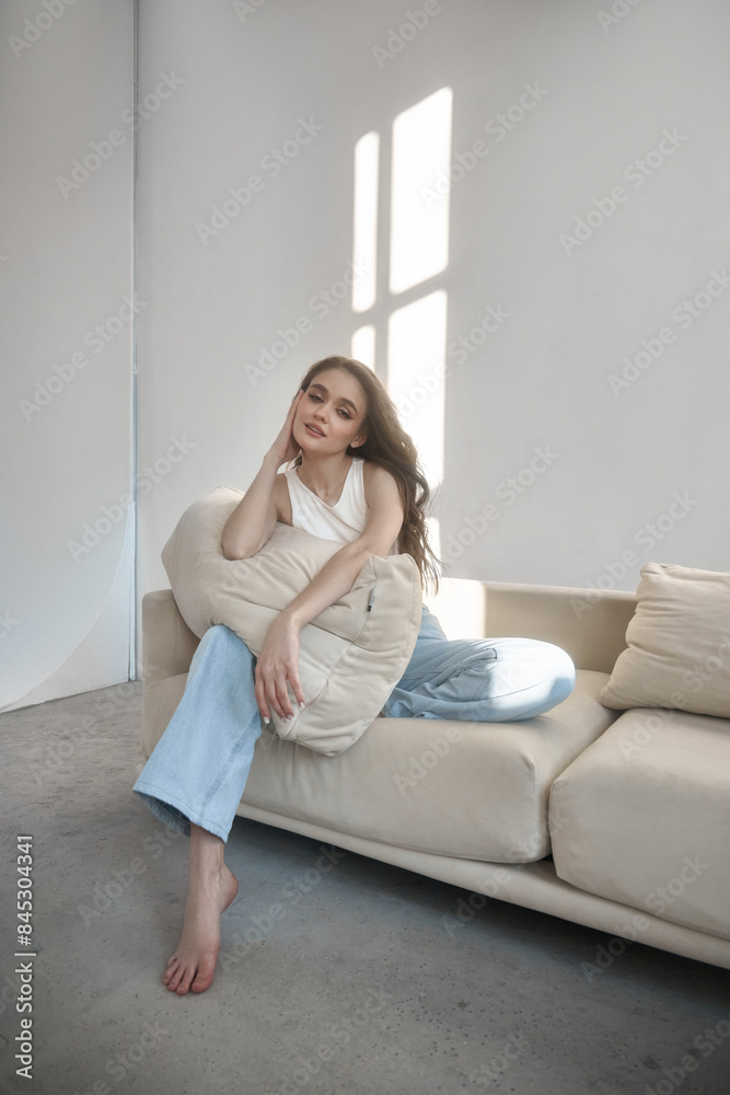 Young beautiful woman with long dark hair sits on white sofa, running her fingers through hair. Room is bright and sunny, with light streaming in through the window Young beautiful woman with long dark hair sits on white sofa, running her fingers through hair. Room is bright and sunny, with light streaming in through the window
