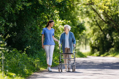 Female caregiver and senior woman with walker on walk in nature. Nurse and elderly woman enjoying a warm day in nursing home, public park. © Halfpoint