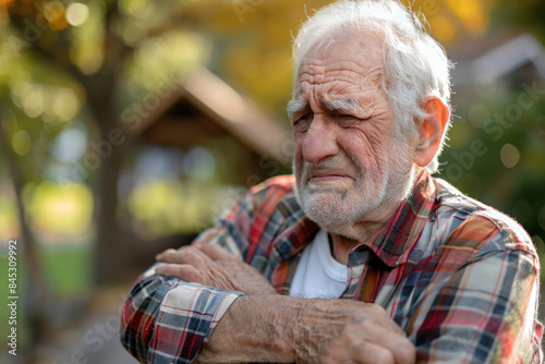An elderly man stands outdoors, wearing a plaid shirt, with his arms crossed, looking somber