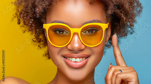 Portrait of a happy young woman wearing yellow sunglasses and pointing her finger to her temple with a big smile