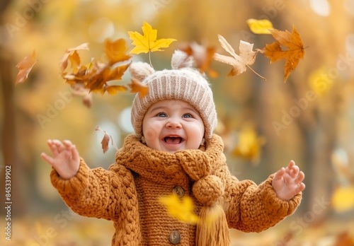 Cute happy smiling baby playing with autumn leaves on the ground, in a park, with yellow trees in the background, depicting the fall season concept.