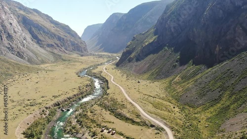 A smooth flight over a narrow mountain valley between mountains. An amazing green river with rapids. Bright daytime sun.