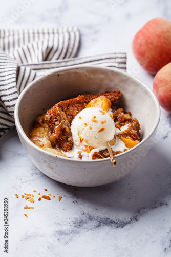 Portion of peach cobbler with scoop of ice cream in bowl, white marble background.