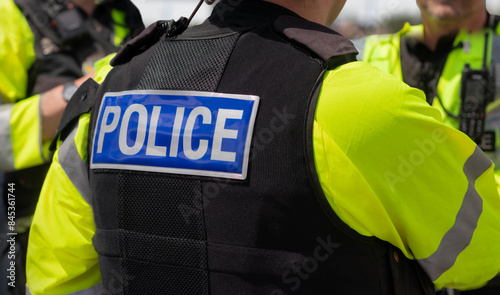 Police - Close-up of 'POLICE' marking on the back of a hi-visibility stab proof vest worn by a trio of police officers at the scene of an incident. Officers enforce the law and establish public order.