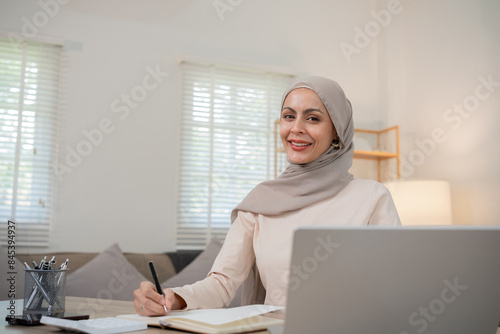 Confident Muslim Woman Working from Home, Wearing Hijab, Smiling at Desk with Laptop and Notebook in Modern Home Office Setting