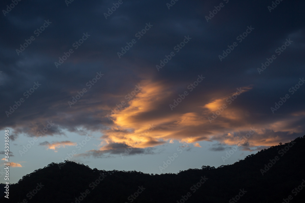 Landscape of thick clouds, orange light hitting the blue sky, silhouettes of trees, forests, and mountains. Area for text in the middle of the image Landscape of thick clouds, orange light hitting the blue sky, silhouettes of trees, forests, and mountains. Area for text in the middle of the image