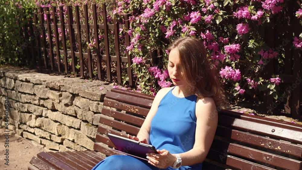Woman in blue dress working on tablet online on remote job outdoor. Business lady sitting on a bench and typing on pad in the park. Modern workplace in the city. Urban lifestyle Woman in blue dress working on tablet online on remote job outdoor. Business lady sitting on a bench and typing on pad in the park. Modern workplace in the city. Urban lifestyle