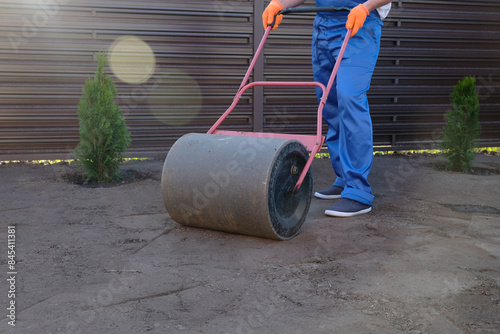 male worker in a uniform prepares a plot of land for sowing a lawn using a garden roller, leveling and compacting the earth