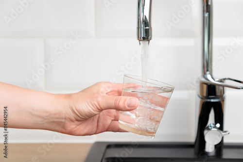 Thirst human wanted to drink water and filling up a glass of tap pure water from kitchen faucet with white tiles wall. Concept of purity and cleanness of fresh natural water, banner with copy space
