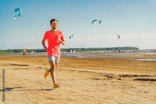 A man in a bright outfit runs along a sandy beach under a clear sky with colorful kites flying above.