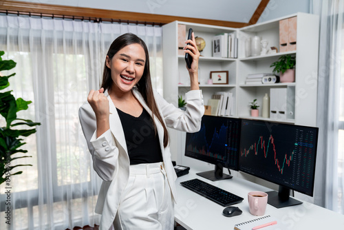 Successful young Asian businesswoman looking at camera with smile face, earning high profit raising fist up, holding smartphone in dynamic exchange stock market graph at modern office. Stratagem. photo