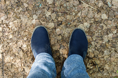 Men's legs in blue jeans and slip-on shoes on the ground covered with leaves, first-person view