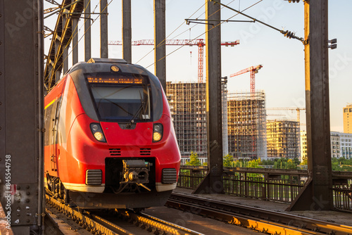 Electric commuter train on a steel railway bridge over a river at sunset in summer