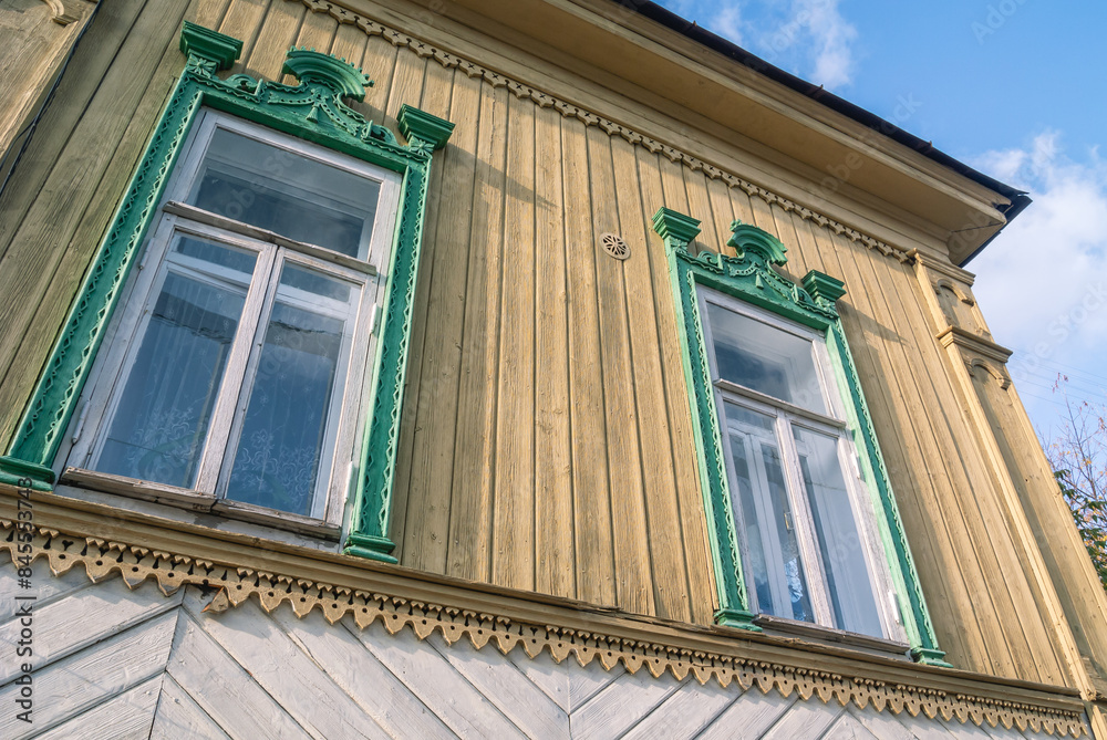 Obraz premium Windows with carved wooden frames of an old log house, sheathed in boards. An old wooden house and a blue sky. Architecture and traditions in the last century.