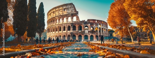 A large Roman colosseum is surrounded by trees and people. The leaves on the ground create a beautiful autumn scene