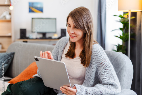 Attractive woman, freelancer holding digital tablet, using, ordering, sitting on comfortable sofa