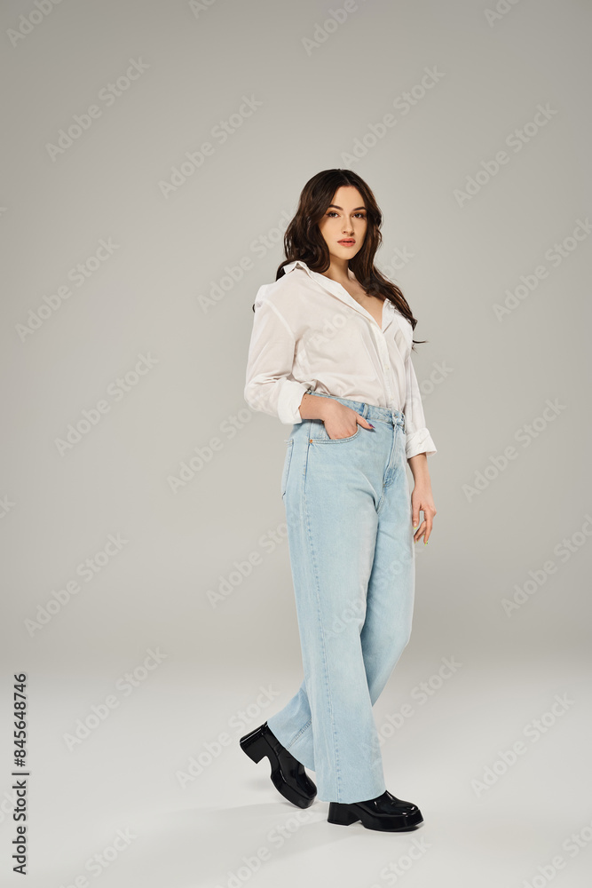 Stylish plus size woman posing confidently in white shirt and blue jeans on a gray backdrop. Stylish plus size woman posing confidently in white shirt and blue jeans on a gray backdrop.