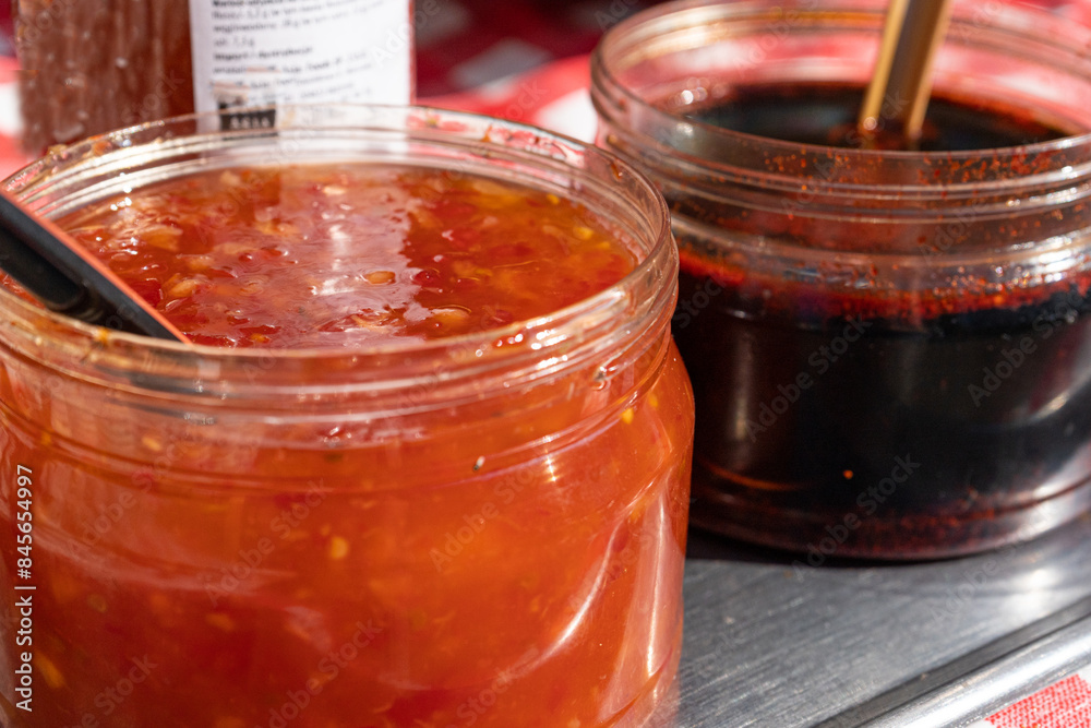 Two jars with red and dark sauces on a metal table, with spoons. Chunky, spicy red sauce and dark Two jars with red and dark sauces on a metal table, with spoons. Chunky, spicy red sauce and dark