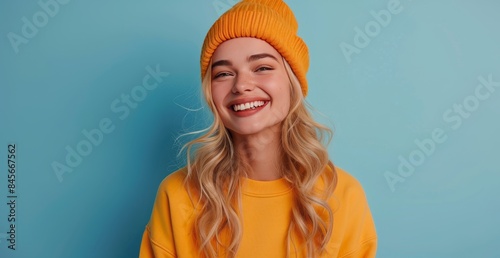 Smiling Young Woman Wearing a Yellow Hoodie and Beanie Against a Blue Background