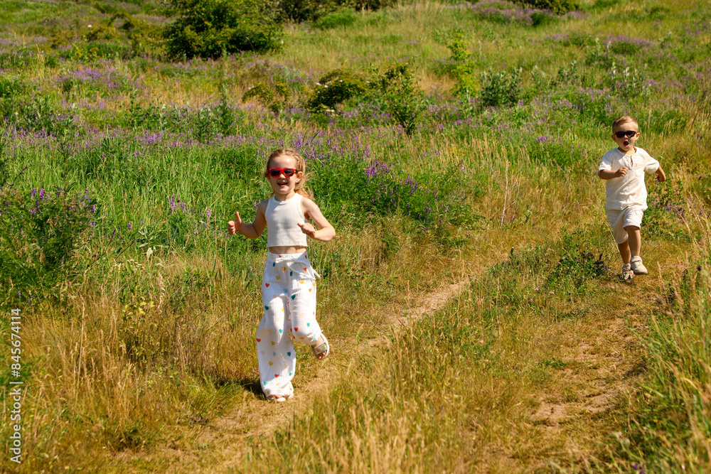 A little boy and girl in sunglasses run happily along a path in a flowering meadow in summer A little boy and girl in sunglasses run happily along a path in a flowering meadow in summer