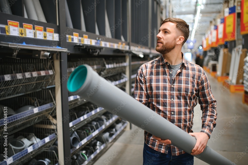 Naklejka premium Handsome workman choosing sewer pipes standing near the showcase of the plumbing shop