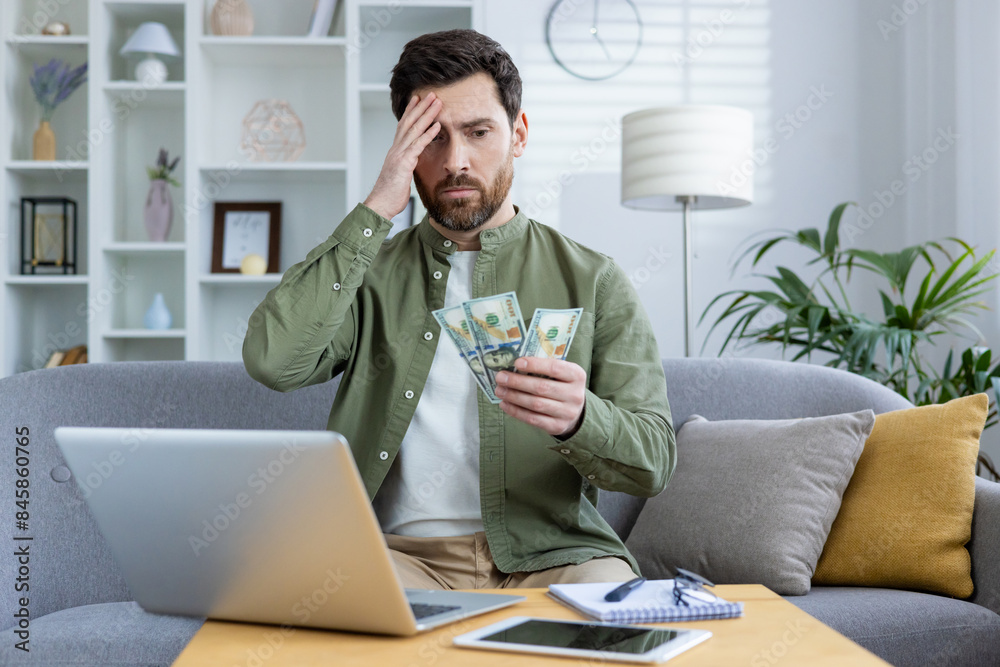 Worried young man sitting at home on the sofa in front of the table with a laptop, holding his head with his hand and looking sadly at the cash money in his hand. Worried young man sitting at home on the sofa in front of the table with a laptop, holding his head with his hand and looking sadly at the cash money in his hand.