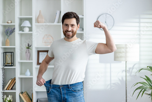 фотографія Portrait of a happy and confident young man at home, showing his weight and waist in oversize jeans, showing a sit and victory gesture with his hand