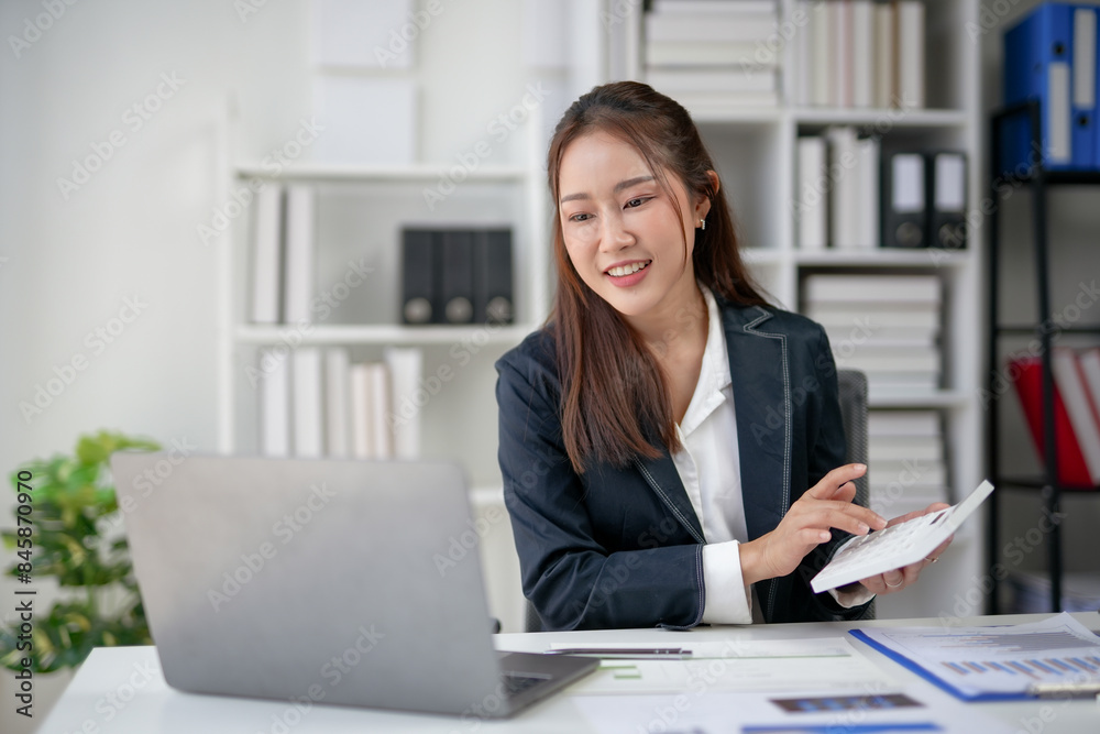 Businesswoman working on a laptop in a modern office, holding a notepad, and smiling. Professional work environment with shelves and documents. Businesswoman working on a laptop in a modern office, holding a notepad, and smiling. Professional work environment with shelves and documents.