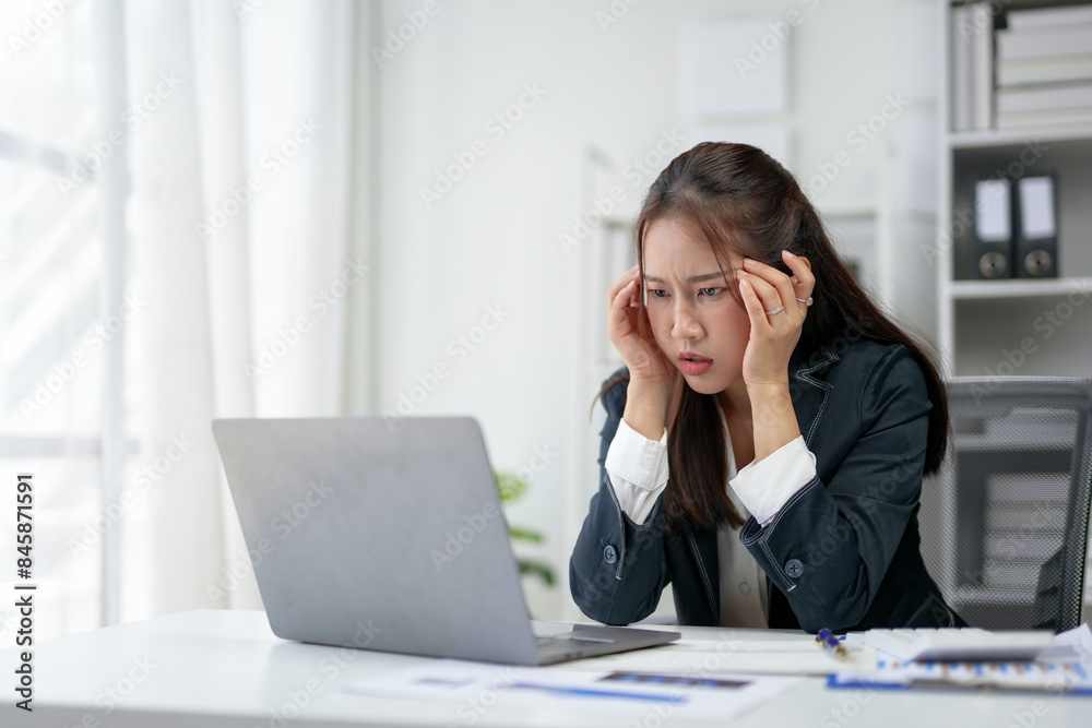 Young businesswoman frustrated at her desk, staring at a laptop screen. Office environment with documents and files in the background. Young businesswoman frustrated at her desk, staring at a laptop screen. Office environment with documents and files in the background.