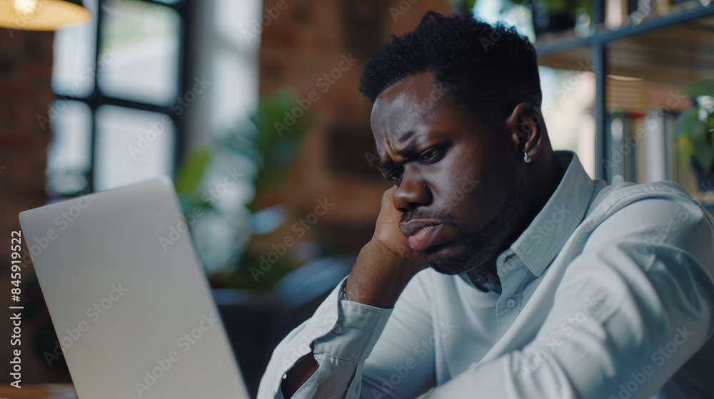 Obraz premium A man appears deeply engaged in his laptop screen, with a furrowed brow and hand on cheek, set in a modern, cozy office space.
