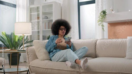 African American woman girl lady user influencer female come in living room walking going in home sit relax at couch sofa using mobile phone internet addict social media smartphone addiction chatting