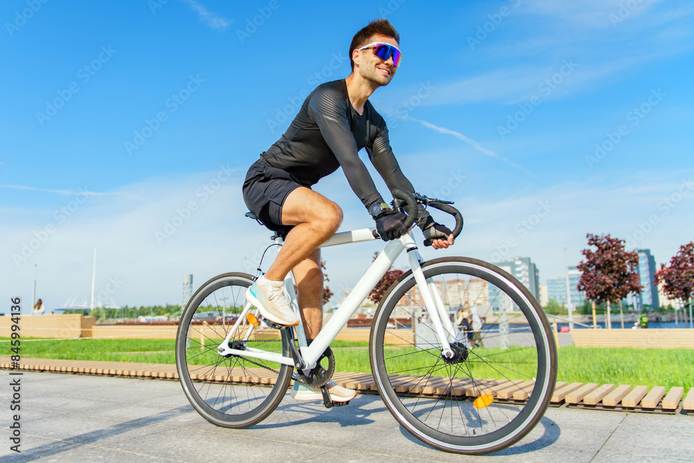 Naklejka premium A man in black cycling attire rides a white bike on a sunny day, smiling, with urban greenery in the background.