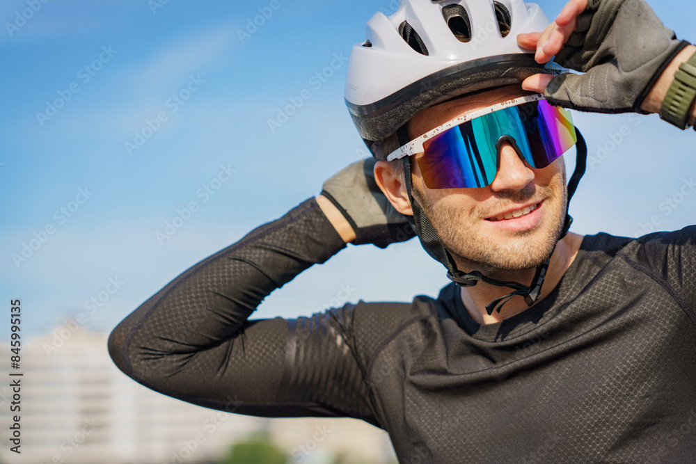 A cyclist in black attire adjusts his white helmet while wearing reflective sunglasses, smiling under a clear sky. A cyclist in black attire adjusts his white helmet while wearing reflective sunglasses, smiling under a clear sky.