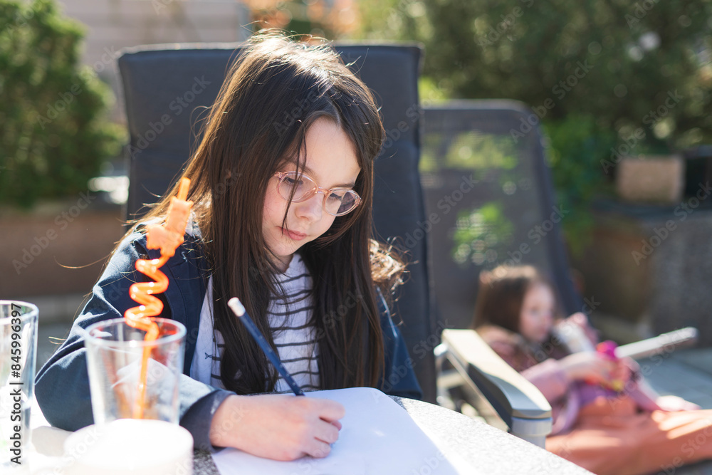 Fototapeta premium A little girl with glasses sits at a table in the backyard and does her homework. Educational concept