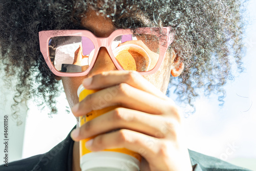 Young mixed-race non-binary person with an afro and sunglasses, wearing a black business suit. Drinking coffee while the reflection is visible in the sunglasses.