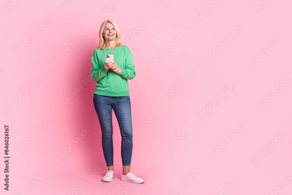 Full length photo of lovely senior lady look empty space hold device dressed stylish green garment isolated on pink color background Full length photo of lovely senior lady look empty space hold device dressed stylish green garment isolated on pink color background