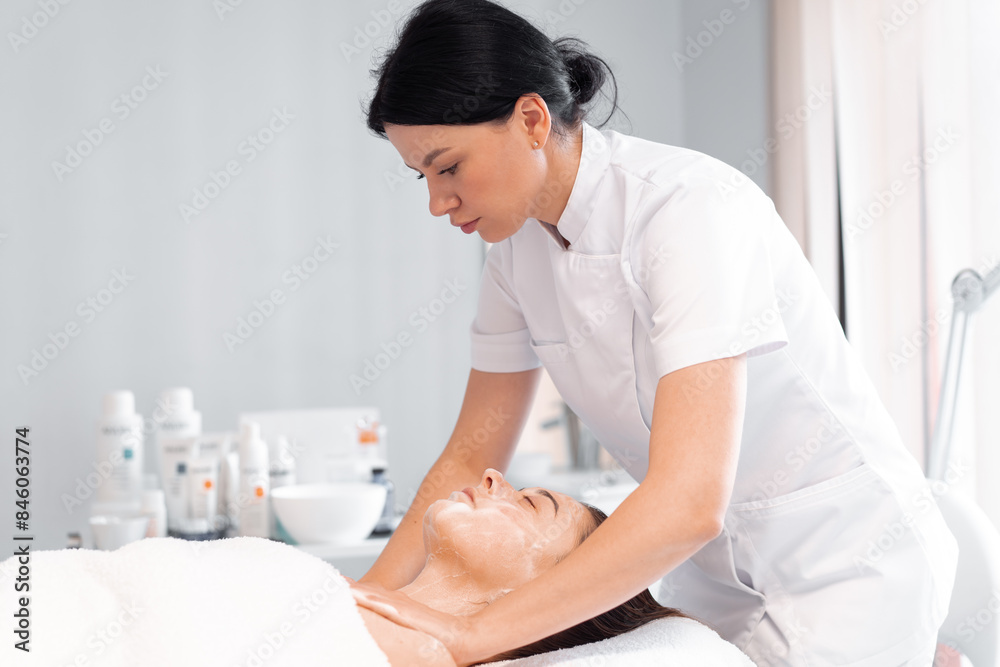 Female cosmetologist in uniform cleanses the skin of woman with foam. Woman's skin care procedure in beauty clinic. Natural cosmetics. Cosmetic treatment of face. Therapeutic peeling cream. Female cosmetologist in uniform cleanses the skin of woman with foam. Woman's skin care procedure in beauty clinic. Natural cosmetics. Cosmetic treatment of face. Therapeutic peeling cream.