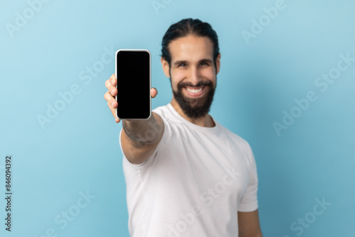 Portrait of positive man with beard wearing white T-shirt standing holding phone with empty display, looking at camera with toothy smile. Indoor studio shot isolated on blue background.
