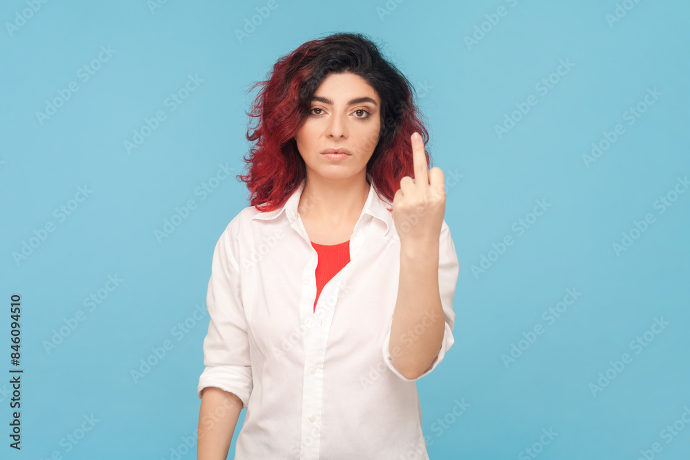 Portrait of rude impolite woman with fancy red hair showing middle finger, arguing with her husband, being dissatisfied, wearing white shirt. Indoor studio shot isolated on blue background. Portrait of rude impolite woman with fancy red hair showing middle finger, arguing with her husband, being dissatisfied, wearing white shirt. Indoor studio shot isolated on blue background.