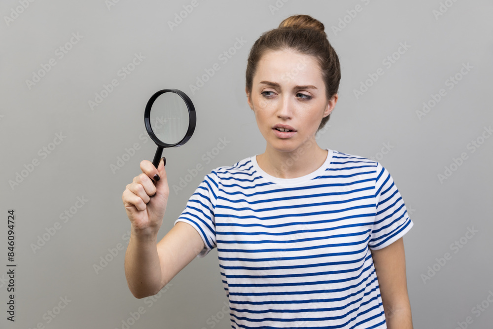 Portrait of woman wearing striped T-shirt looking through magnifying glass, finding out something, magnifies small details to see, inspecting. Indoor studio shot isolated on gray background. Portrait of woman wearing striped T-shirt looking through magnifying glass, finding out something, magnifies small details to see, inspecting. Indoor studio shot isolated on gray background.