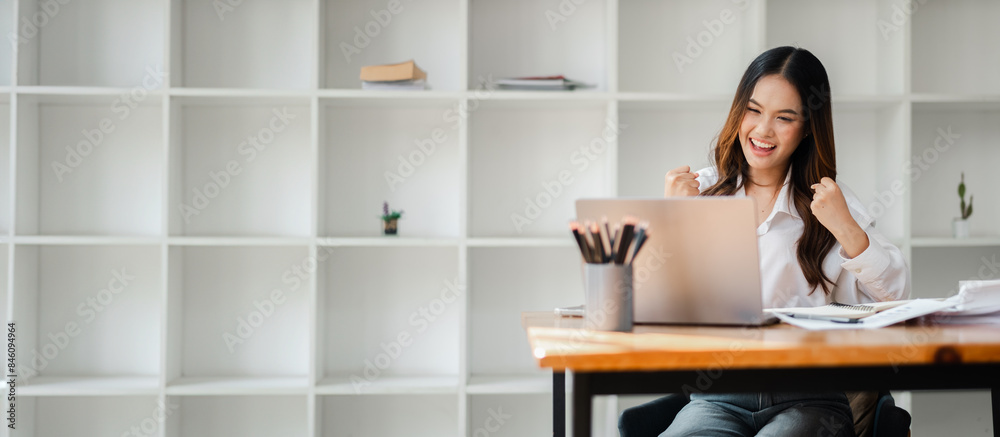 Excited young woman celebrating success while working on a laptop in a modern office with minimalist white shelves in the background. Excited young woman celebrating success while working on a laptop in a modern office with minimalist white shelves in the background.