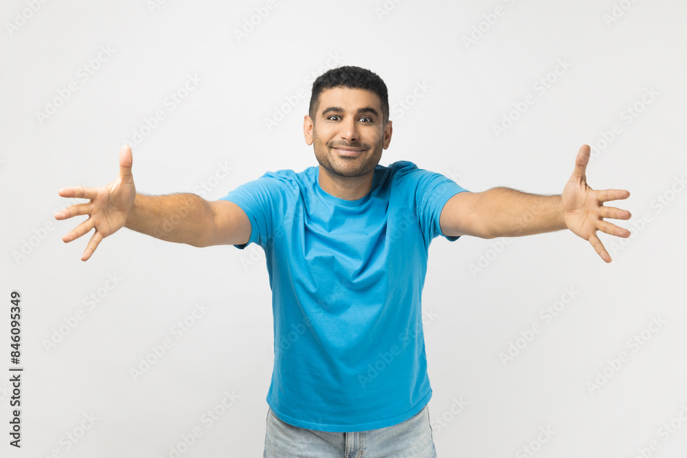 Portrait of friendly cheerful positive unshaven man wearing blue T- shirt standing spread arms, inviting to hug, looking at camera with toothy smile. Indoor studio shot isolated on gray background.