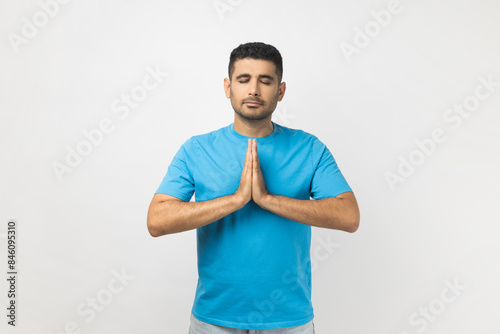 Portrait of calm attractive young adult unshaven man wearing blue T- shirt standing practicing yoga, keeps eyes closed, trying to relax. Indoor studio shot isolated on gray background. © khosrork