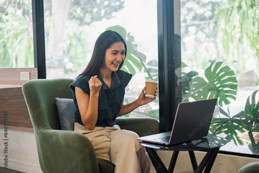 A young woman celebrates success while working remotely on her laptop in a modern home office with a lush greenery background.